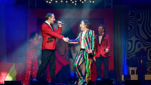 CIRQUE - The Greatest Show cast on stage with a man in a rainbow suit looking surprised as he shakes the hand of a man in a red sequin jacket who is singing to him
