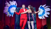CIRQUE - The Greatest Show performers posing including a man in a red suit winking and a lady dressed in a black and pink dress with pom poms hugging him