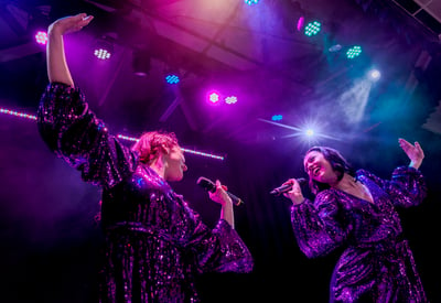 Queens of the Dancefloor - two of the actresses sing to each other with smiles on their faces