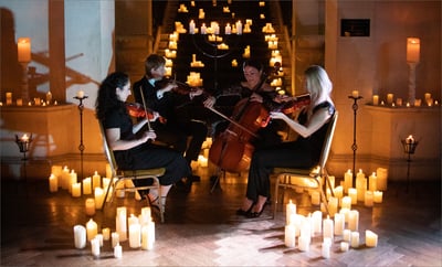 Four people playing violins in a candlelit setting, surrounded by flickering candles and a warm, inviting atmosphere.