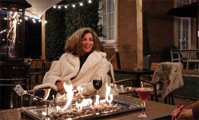 A woman in a fur coat sits at a table by a warm fire, exuding a festive atmosphere and elegance.