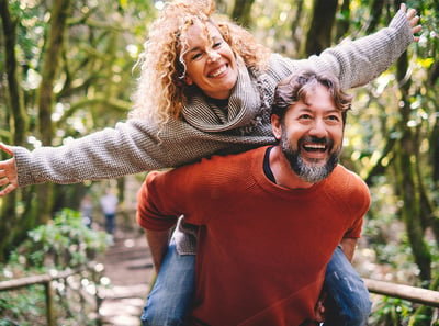 A couple, where the man has the woman on his back and she pretends to fly, both smiling gleefully