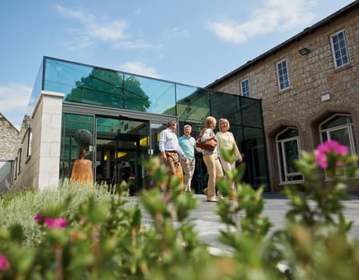 Four people walk towards a modern glass entrance, surrounded by greenery and flowers on a sunny day.