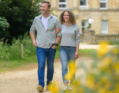 A couple strolls arm in arm along a gravel path, surrounded by lush greenery and colorful flowers, with an elegant building in the background.