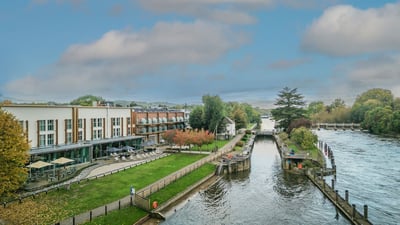 The Runnymede on Thames Hotel by a tranquil river, surrounded by trees and buildings, creating a picturesque landscape.