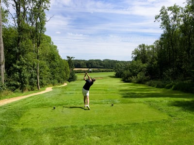 A golfer takes a shot on the scenic Heythrop Park course, surrounded by vibrant greenery and a clear blue sky.