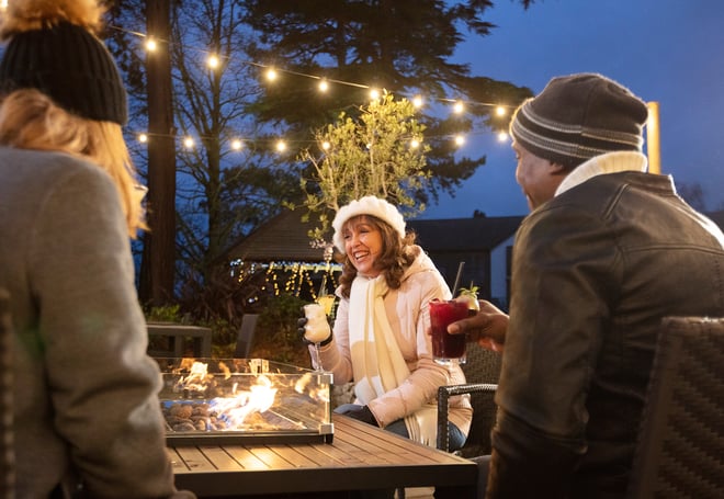 A guest laughs joyfully as she's surrounded by friends beside a firepit on the terrace at Alvaston Hall