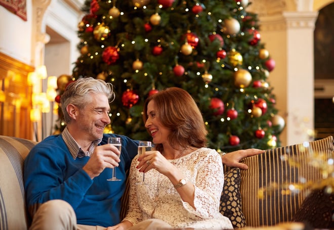 A couple enjoying a drink together in front of a Christmas tree and smiling at one another in the Great Hall at Thoresby Hall