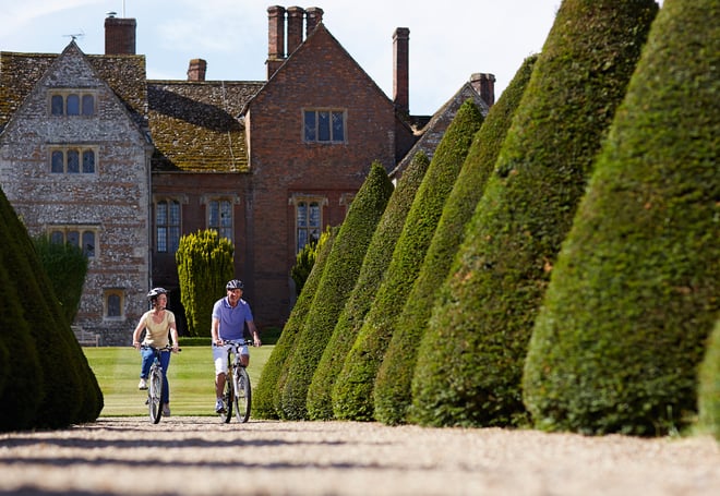 A couple cycling between the large shrubs in the foreground in front of Littlecote House Hotel