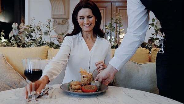 A woman in a white blouse sits at a table, reaching for a plate of food as a waiter serves her, with a glass of red wine nearby.