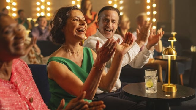 A group of audience members applauding enthusiastically with drinks on tables in a lively, warmly lit setting.