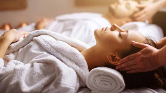 A relaxing spa scene with two individuals lying down, wrapped in white towels, enjoying head massages in a tranquil environment.