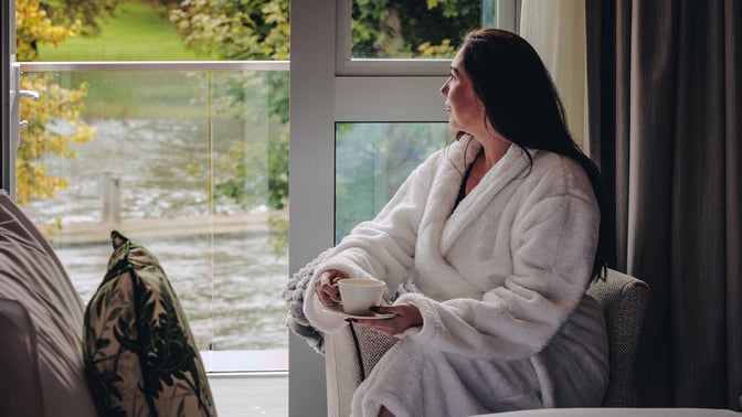 A woman in a robe sits comfortably in a chair, gazing out the window, bathed in soft natural light.