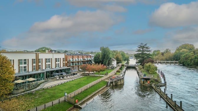 The Runnymede on Thames Hotel by a tranquil river, surrounded by trees and buildings, creating a picturesque landscape.